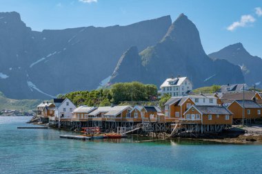 Norveç 'te sakin bir fiyordun kenarında, arka planda yükselen dağlarla birlikte renkli evlerden oluşan antika bir köy bulunuyor. Sarı balıkçı Rorbuer kulübeleri, Sakrisoy, Lofoten, Norveç