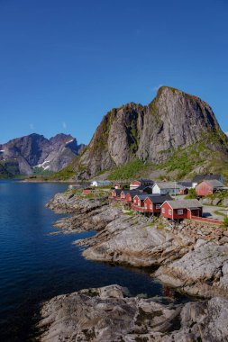 Norveç 'te, yüksek dağlarla çevrili kayalık kıyı şeridine tünemiş kırmızı kulübeleri olan küçük bir köy manzarası. Lofoten Adaları 'ndaki Hamnoy balıkçı köyü, Norveç' te kırmızı rorbu evleri var.