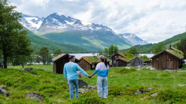 A couple walks hand in hand in front of traditional Norwegian cabins with a stunning mountain backdrop. Innerdalen Norway most beautiful mountain valley clipart