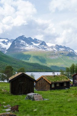 İki geleneksel Norveç kulübesi, manzaralı bir göl ve uzaktaki karla kaplı dağlara bakan çimenli bir tarlada duruyor. Trollheimen dağlarının iç kesimlerindeki vadi