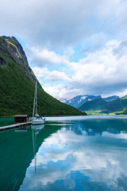 A white sailboat is docked at a wooden pier in a calm fjord. The mountains are reflected in the still water. Norangsfjorden Norangdal Urke Norway clipart