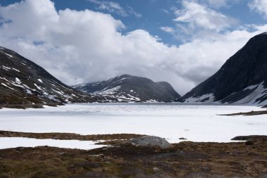 Norveç dağlarında donmuş bir göl, karla kaplı tepelerle çevrili. Gökyüzü mavi ve beyaz bulutların bir karışımıdır. Langvatnet Geiranger Norveç