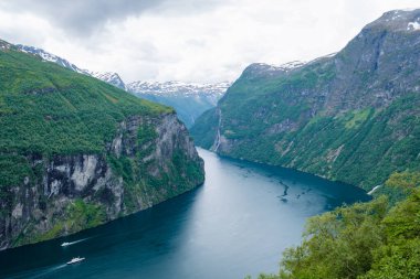 Etrafı yemyeşil dağlarla çevrili bir Norveç fiyordunun nefes kesici manzarası. Su, bulutlu gökyüzünü yansıtan koyu mavi bir renktir. Norveç 'in Geiranger' si