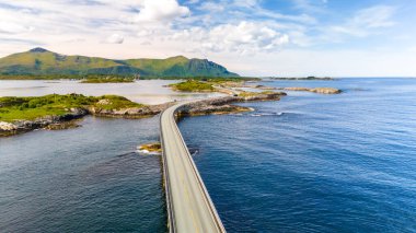 Norveç adaları boyunca uzanan temiz mavi su ve arka planda yeşil tepelerle çevrili bir yolun havadan görüntüsü. Atlantik Okyanusu Yolu Norveç