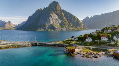Sahil şeridi boyunca uzanan canlı mavi suları, yemyeşil dağları ve büyüleyici evleri olan huzurlu Norveç fiyordunun resimsel bir görüntüsü. Reine, Lofoten, Norveç 