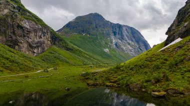 Norveç 'teki bir vadi manzarası. Etrafındaki dağları ve bulutlu gökyüzünü yansıtan sakin bir göl sergileniyor..