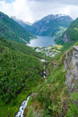 Norveç 'te yemyeşil bir dağ yamacından dökülen bir şelalenin manzarası, bir fiyort ve uzaktaki bir köy. Norveç 'in Geiranger' si