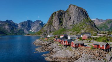 Norveç 'te bir fiyordun kayalık kıyılarındaki kırmızı kulübelerin manzarası. Etrafı yüksek dağlarla çevrili, Lofoten Adaları 'ndaki Hamnoy balıkçı köyü, Norveç' te kırmızı rorbu evleriyle çevrili.