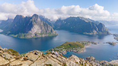 Norveç 'te Reine, dağların, fiyortların ve adaların göz kamaştırıcı manzarasına bakan bir uçurumda oturan bir çift. Reinebringen, Lofoten, Norveç 'te bir dağın tepesinde bir çift.