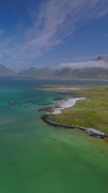 Turkuaz suların, yeşil çayırların ve mavi gökyüzünün altında görkemli dağların bulunduğu güzel bir kıyı manzarası. Lofoten, Norveç