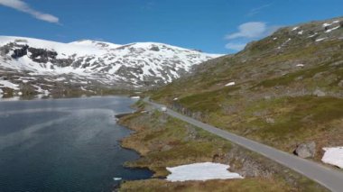 Huzurlu bir dağ yolu, karlı zirveler ve yemyeşil yollarla çerçevelenmiş sakin bir gölün yanında esiyor. Langvatnet, Geiranger, Norveç