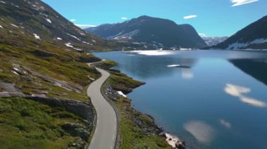 Sakin bir göl kıyısında, karlı tepeleri olan bir dağ yolu huzur veren manzarayı nefes kesici bir şekilde sunar. Langvatnet, Geiranger, Norveç