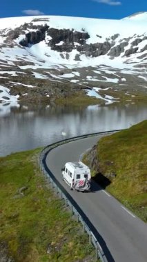 Dolambaçlı bir dağ yolunda, gölü ve karla kaplı tepeleri olan bir karavanın çarpıcı manzarası. Langvatnet, Geiranger, Norveç