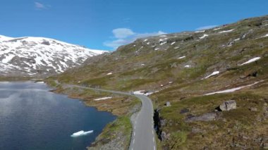 Norveç 'in pitoresk dağ yolu baharda devam eden kar yamaçları olan sakin bir gölün yanında kıvrılıyor. Langvatnet, Geiranger, Norveç