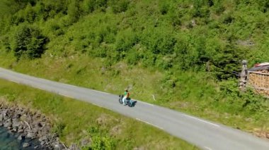 Güneşli bir günde, verimli ormanlar ve berrak mavi sularla çevrili bir sahil yolunda bisiklet süren huzurlu bir kadın. Lovatnet, Norveç