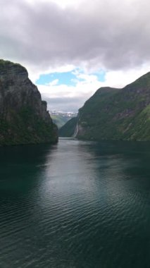 Şelaleli güzel fiyort manzarası, yemyeşil dağlar ve bulutlu bir gökyüzünün altında sakin bir su kütlesi. Geiranger, Norveç