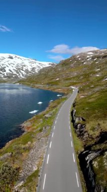 Arka planda karla kaplı zirveleri olan bir dağ gölünün etrafında dönen manzaralı bir yol manzarası. Langvatnet, Geiranger, Norveç