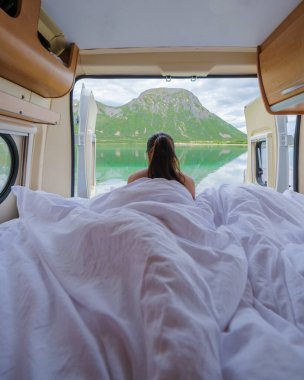 Bir kadın bir karavanda uyanıyor, pencere Norveç dağı ve gölünün çarpıcı manzarasını gösteriyor. Lofoten Norveç