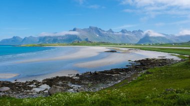 Geniş, kumlu bir sahil mavi bir gökyüzünün altında uzanır, yeşil çayırlar ve sisle çevrili dağlar. Lofoten Norveç