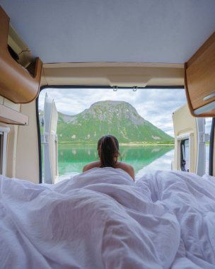 Norveç 'te muhteşem bir dağ gölü manzaralı bir karavanda uyanan bir kadın. Sabah güneşi sahneyi altın bir ışıkla aydınlatıyor. Lofoten Norveç