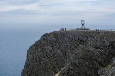Bir grup turist, Norveç 'in Kuzey Burnu' ndaki ikonik küresel anıta hayran kalarak bir uçurumun kenarında duruyorlar. Uçsuz bucaksız okyanus önlerinde uzanır bulutlu bir gökyüzünün altında..