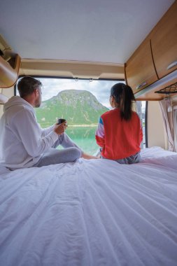 Bir çift, Norveç 'te yeşil bir dağ ve göl manzaralı bir karavanda dinlenme sabahının tadını çıkarıyor. Asyalı kadınlar ve Avrupalı erkekler Lofoten Norveç 'te bir karavanda