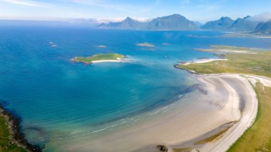 Norveç 'te açık mavi su ve yeşil adalarla çevrili uzun ve kumlu bir sahil manzarası. Kumsal yavaşça denize doğru kıvrılır ve uzaktaki tepeler yuvarlanır. Kolbedeliden Sahili, Lofoten