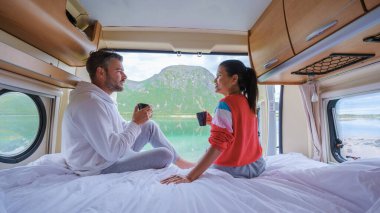 Bir çift, kamp minibüslerinde bir fincan kahvenin keyfini çıkarıyor. Norveç dağlarının muhteşem manzarası ve dışarıda bir göl var. Lofoten Norveç