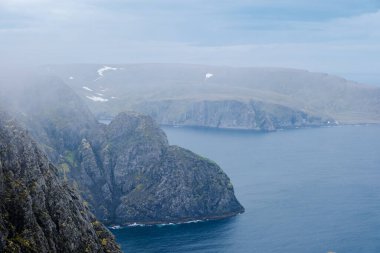 Engebeli kayalıklardan oluşan puslu bir manzara ve engin mavi deniz Kuzey Norveç 'te puslu bir gökyüzünün altında uzanıyor. Nordkapp, Kuzey Burnu
