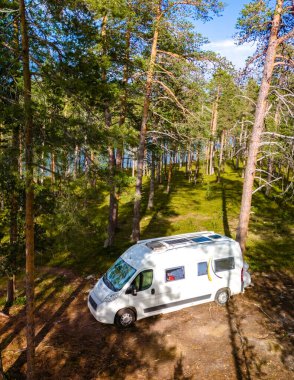 Finlandiya 'nın Laponya ormanında yüksek çam ağaçlarıyla çevrili bir açıklığa beyaz bir karavan park edilmiştir. Minibüs, ağaçların arasından süzülen güneş ışığıyla birlikte toprak bir yolda duruyor..