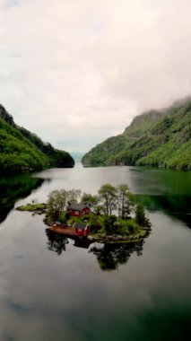 Arkanıza yaslanın ve sakin bir adanın göldeki iki büyüleyici kırmızı evi, yemyeşil ağaçları ve uzaktaki görkemli dağları Lovrafjorden, Norveç 'teki sakin bir videosunu izlerken rahatlayın.