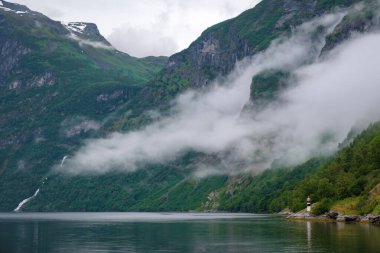 Yalnız bir deniz feneri kayalık bir sahilde duruyor, sakin bir fiyorda tepeden bakan, sisli dağların arasında. Geiranger Fiyordu