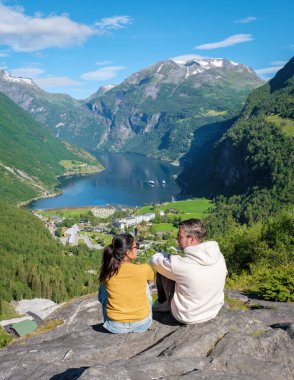Dağlar ve yemyeşil yeşilliklerle çevrili bir Norveç fiyortunun nefes kesici manzarasının tadını çıkaran iki kişi kayalık bir uçurumda oturuyor. Geiranger Fjord Norveç 'te çeşitli kadın ve erkekler var.
