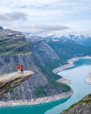 Bir çift, Norveç dağlarındaki dolambaçlı bir turkuaz fiyorda bakarak kayalık bir uçurumun kenarında duruyor. Erkekler ve kadınlar Norveç 'te Trolltunga' da yürüyüş yapıyorlar.