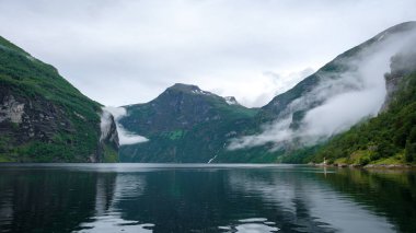 Norveç 'teki dingin bir fiyort, sisle kaplı yüksek uçurumlar tarafından çerçevelenmiş. Sakin sular görkemli manzarayı yansıtıyor. Huzurlu ve hayranlık uyandırıcı bir manzara yaratıyor. Geiranger Fiyordu