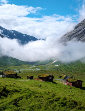 Düşük seviyeli bulutların rüya gibi bir atmosfer yarattığı yemyeşil bir vadide yuva yapmış geleneksel Norveç kulübelerinin resimsel bir sahnesi..