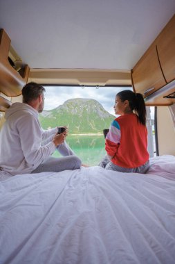 Bir çift sabah kahvelerini Norveç gölü ve dağları manzaralı bir karavanda içiyorlar. Lofoten Norveç 'te bir karavanda bir kadın ve erkek.