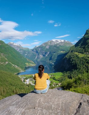 Asyalı bir kadın sersemletici bir Norveç fiyorduna bakan kayalık bir çıkıntıda oturuyor, yemyeşil dağlar ve açık mavi bir gökyüzü ile çevrili. Geiranger Fjord Norveç