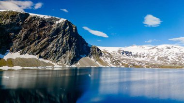 Sakin bir dağ gölü, Norveç 'in engebeli arazisindeki mavi gökyüzünü ve karlı tepeleri yansıtır. Strynefjellsvegen, Geiranger, Norveç