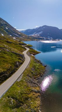 Norveç dağları boyunca kıvrılan ve yanında parlak bir göl bulunan bir yol manzarası. Langvatnet, Geiranger, Norveç