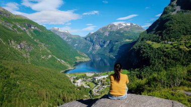 Asyalı bir kadın bir kayanın üzerinde oturmuş, yemyeşil dağlarla çevrili Norveç fiyortunun nefes kesici manzarasına bakıyor. Geiranger Fjord Norveç