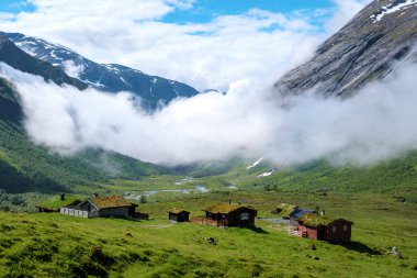 Norveç 'teki bir vadi sisle kaplanmış, yemyeşil tepeler ve karla kaplı dağların arasında yuva yapmış tuhaf ahşap kulübelerle dolu bir manzarayı gözler önüne seriyor..