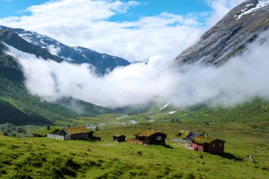 Büyüleyici Norveç vadisi, dağlık kulübeler, yeşil manzaralar, yuvarlanan tepeler, dolambaçlı nehir, uzaklarda bulut toplamak.
