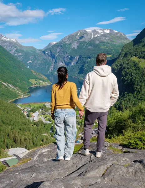 stock image A couple stands hand-in-hand on a rocky cliff overlooking a picturesque Norwegian fjord, surrounded by lush greenery and snow-capped mountains. Geiranger Fjord Norway