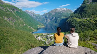 Sersemletici bir Norveç fiyorduna bakan kayalık bir çıkıntıda oturan iki kişi, yemyeşil dağlarla ve açık mavi gökyüzüyle çevrili. Geiranger Fjord Norveç