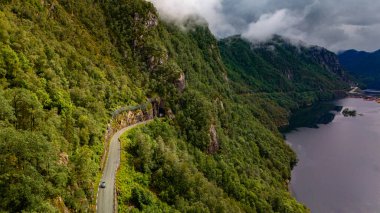 Dolambaçlı bir yol yemyeşil dağların arasından geçiyor, tek bir araba manzaralı geçitten geçiyor. Bulutlar vadinin üzerinde süzülüyor, aşağıdaki mavi sudan yansıyor. Lovrafjorden, Norveç