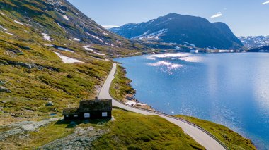 Resimli bir yol yemyeşil dağların arasından geçiyor, el değmemiş mavi bir göle bakıyor, kenarında küçük bir kulübe var. Langvatnet, Geiranger, Norveç