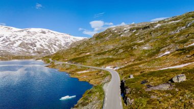 Manzaralı bir yol, mavi gökyüzünü ve karla kaplı zirveleri yansıtan pitoresk bir göl ile nefes kesici Norveç dağları boyunca esiyor. Strynefjellsvegen, Geiranger, Norveç