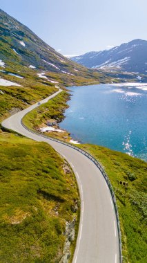 Yeşil dağların arasından geçen dolambaçlı bir yolun ve Norveç 'te parlayan mavi bir gölün havadan görünüşü. Langvatnet, Geiranger, Norveç
