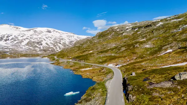 stock image A scenic road winds through a mountainous landscape in Norway. The road hugs the edge of a sparkling blue lake, with snow-capped mountains in the background. Strynefjellsvegen, Geiranger, Norway